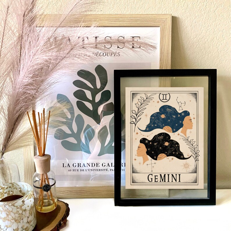 May include: A black framed print with a beige background featuring a zodiac illustration of Gemini. The illustration depicts two women with their hair styled as constellations. The text "II" and "GEMINI" are printed in black.