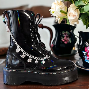 Puede incluir: Botas de plataforma de charol negro con una cadena plateada y abalorios, incluyendo corazones, tazas de té y una tetera. Las botas están sobre una superficie de madera con un jarrón de rosas blancas en el fondo.