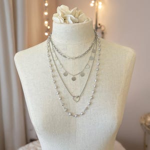 May include: A layered silver-toned necklace set featuring four distinct chains. The top chain is a thick link style, followed by a chain with small circular discs, a chain with a central heart charm, and a long outer chain with small beads.