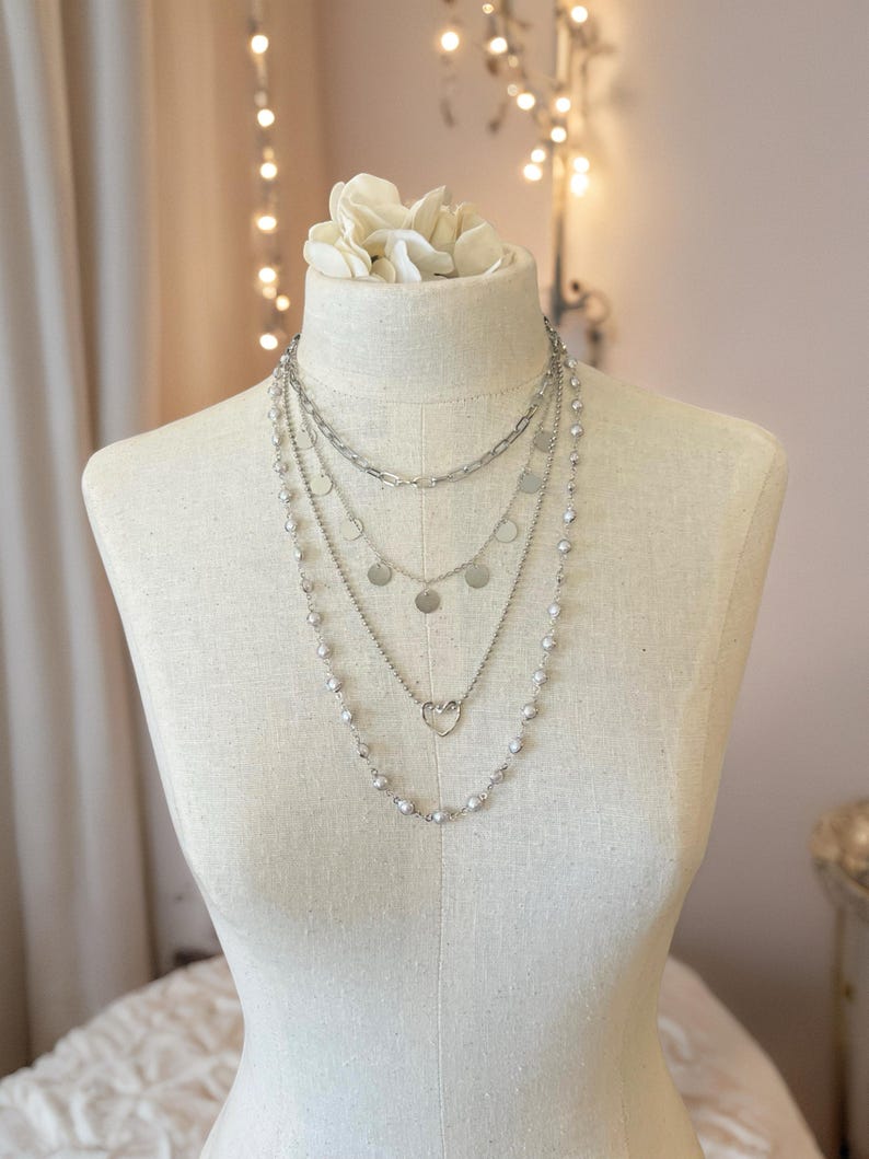 May include: A layered silver-toned necklace set featuring four distinct chains. The top chain is a thick link style, followed by a chain with small circular discs, a chain with a central heart charm, and a long outer chain with small beads.