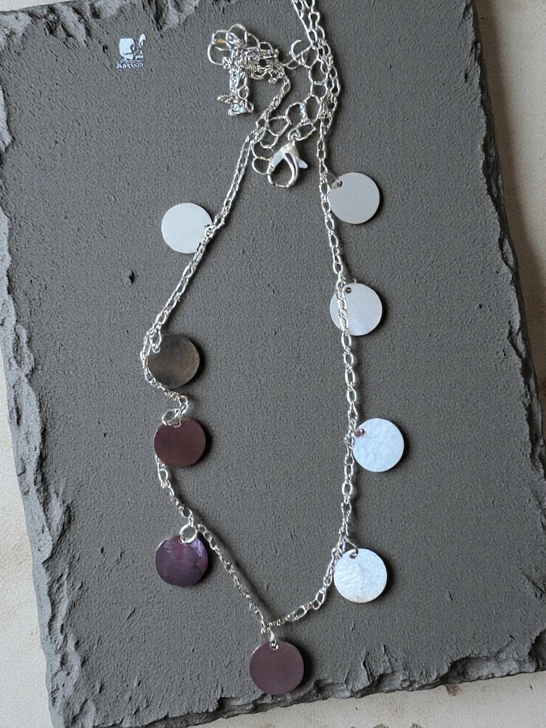 May include: A silver-toned chain necklace featuring multiple small, flat, circular charms in alternating metallic silver and dark brown finishes. The necklace includes a lobster claw clasp and a short extender chain for adjustable length.