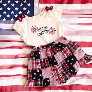 May include: A white t-shirt with red bows on the shoulders and the text "little sparkler" with firework graphics. A red and black plaid skirt with white stars is layered over the shirt. The outfit is laid out on an American flag.