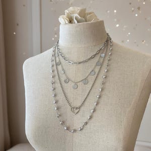 May include: A layered silver-toned necklace set featuring four distinct chains. The top chain is a thick link style, followed by a chain with small circular discs, a chain with a central open heart charm, and an outer chain adorned with small beads.