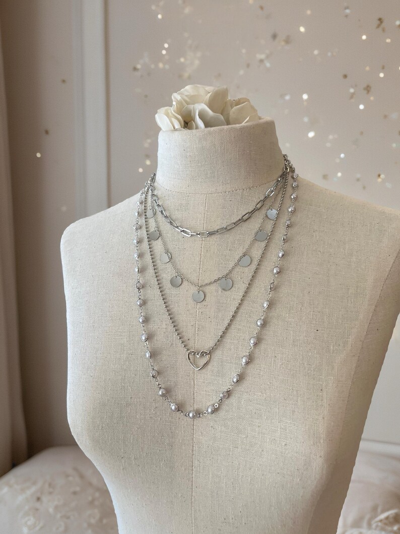 May include: A layered silver-toned necklace set featuring four distinct chains. The top chain is a thick link style, followed by a chain with small circular discs, a chain with a central open heart charm, and an outer chain adorned with small beads.