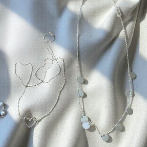 May include: Two silver-toned necklaces. One necklace features a delicate chain with a central open heart-shaped pendant. The second necklace consists of a fine chain adorned with multiple small, flat, circular silver-toned discs spaced evenly along the length.