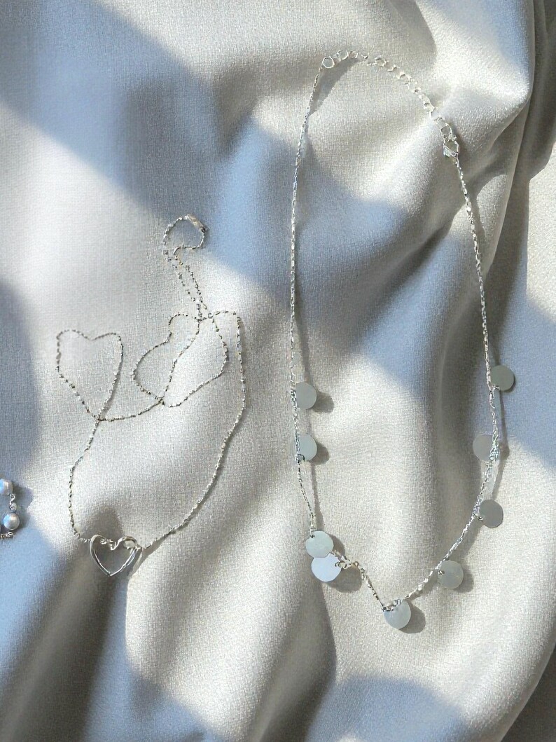 May include: Two silver-toned necklaces. One necklace features a delicate chain with a central open heart-shaped pendant. The second necklace consists of a fine chain adorned with multiple small, flat, circular silver-toned discs spaced evenly along the length.