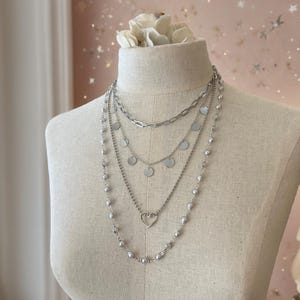 May include: A layered silver-toned necklace set featuring four distinct chains. The top chain is a thick link style, followed by a chain with small circular discs, a delicate chain with a central open heart charm, and a long chain with spaced beads.