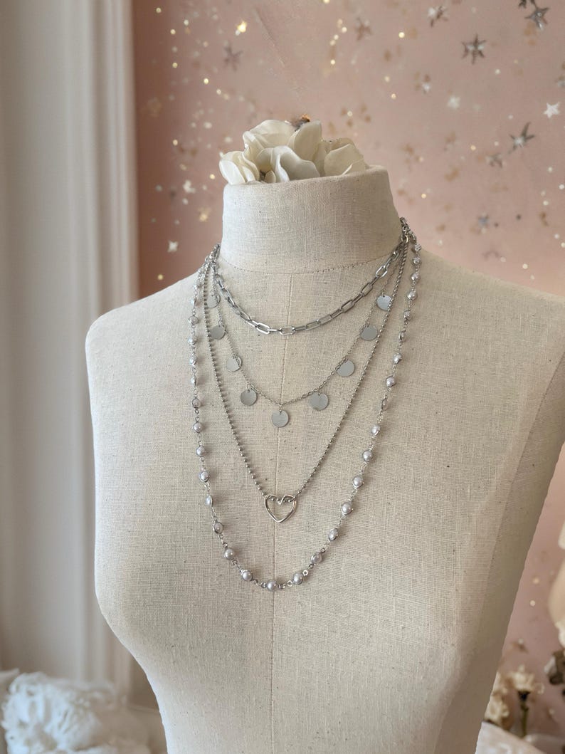 May include: A layered silver-toned necklace set featuring four distinct chains. The top chain is a thick link style, followed by a chain with small circular discs, a delicate chain with a central open heart charm, and a long chain with spaced beads.