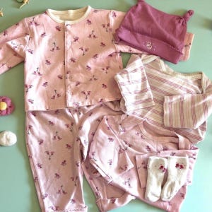 May include: A collection of baby clothing in shades of pink, including a button-up shirt, pants, a long-sleeved onesie, a striped shirt, a beanie, and socks. The pink items feature a floral pattern. The socks are white with pink floral accents.