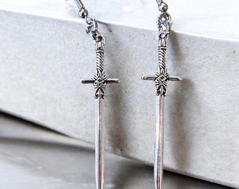 May include: A pair of silver-colored sword earrings. Each earring features a detailed sword design with a cross-shaped hilt and a long, pointed blade. The earrings have a hook closure.
