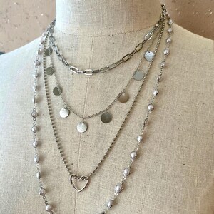 May include: A multi-layered silver-toned necklace set featuring four distinct strands. The layers include a thick rectangular link chain, a delicate chain with small circular metal discs, a thin chain with a heart pendant, and a long beaded strand.
