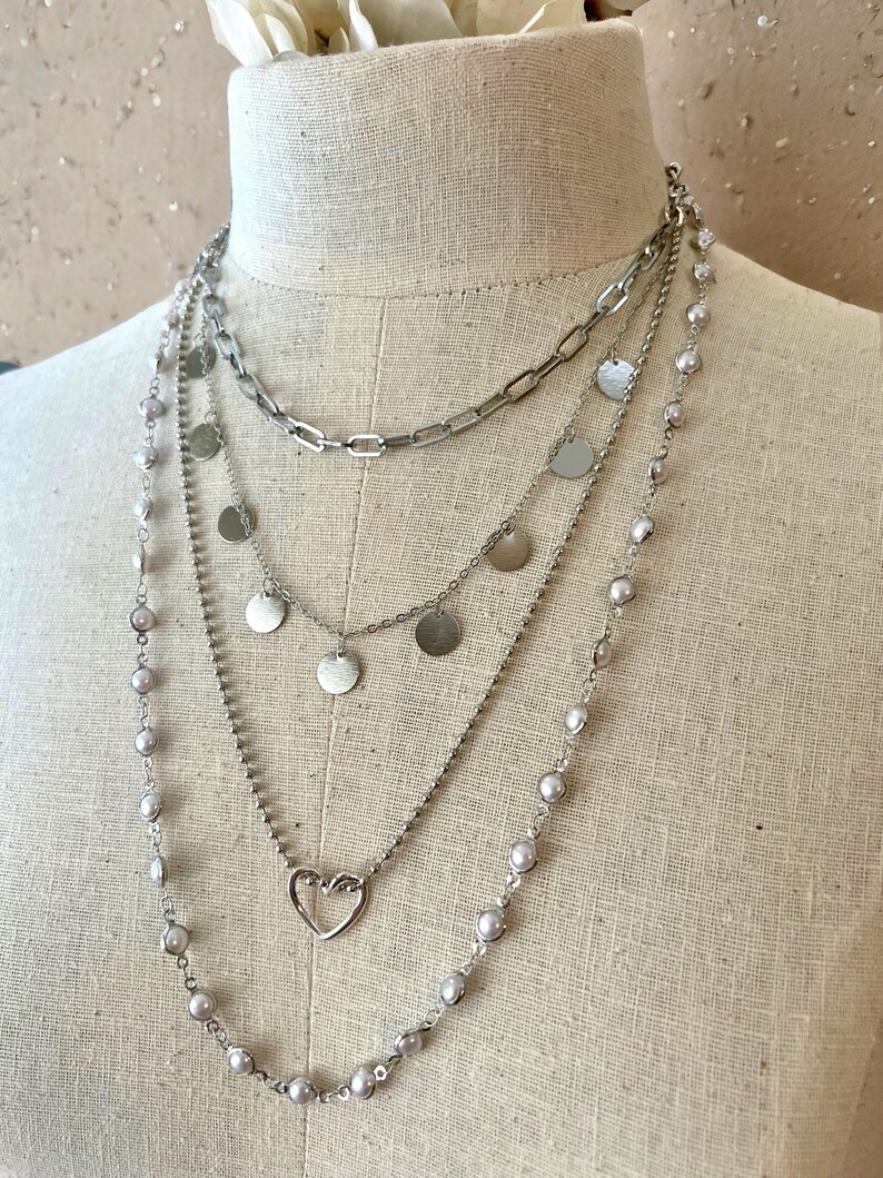 May include: A multi-layered silver-toned necklace set featuring four distinct strands. The layers include a thick rectangular link chain, a delicate chain with small circular metal discs, a thin chain with a heart pendant, and a long beaded strand.