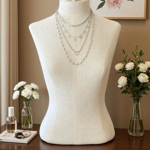 May include: A layered silver-tone necklace set displayed on a beige fabric mannequin. The set features four distinct chains, including one with small circular charms, one with a heart-shaped pendant, and one accented with small white beads.