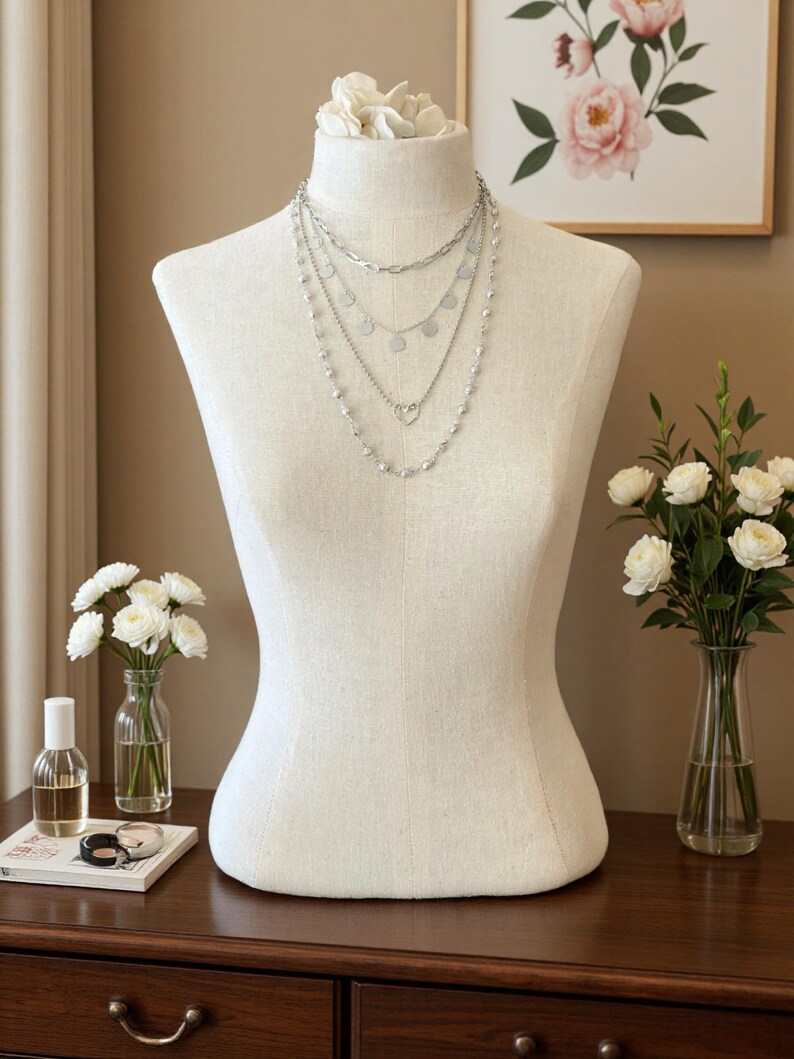 May include: A layered silver-tone necklace set displayed on a beige fabric mannequin. The set features four distinct chains, including one with small circular charms, one with a heart-shaped pendant, and one accented with small white beads.