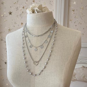May include: A multi-strand silver-toned necklace set featuring four distinct layers. The layers include a thick oval link chain, a delicate chain with small circular discs, a thin chain with a heart-shaped pendant, and a long chain with small beads.