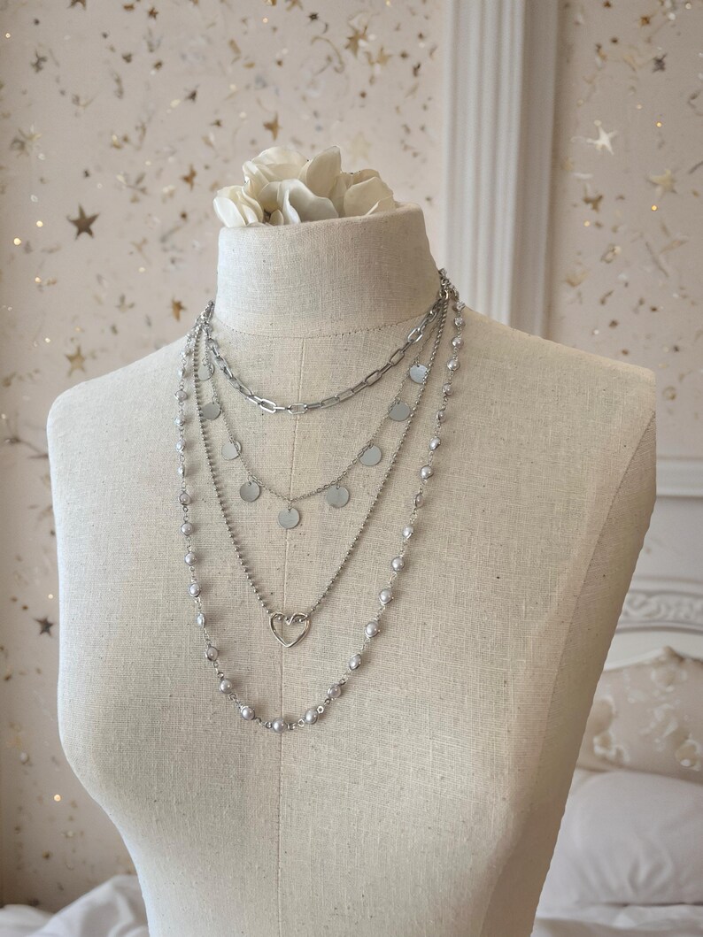 May include: A multi-strand silver-toned necklace set featuring four distinct layers. The layers include a thick oval link chain, a delicate chain with small circular discs, a thin chain with a heart-shaped pendant, and a long chain with small beads.