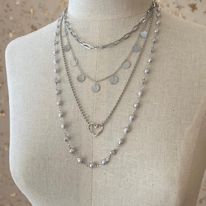 May include: A layered silver-tone necklace set featuring four distinct strands. The top layer is a thick chain, the second has small circular discs, the third features a delicate heart charm, and the longest strand is adorned with small round beads.