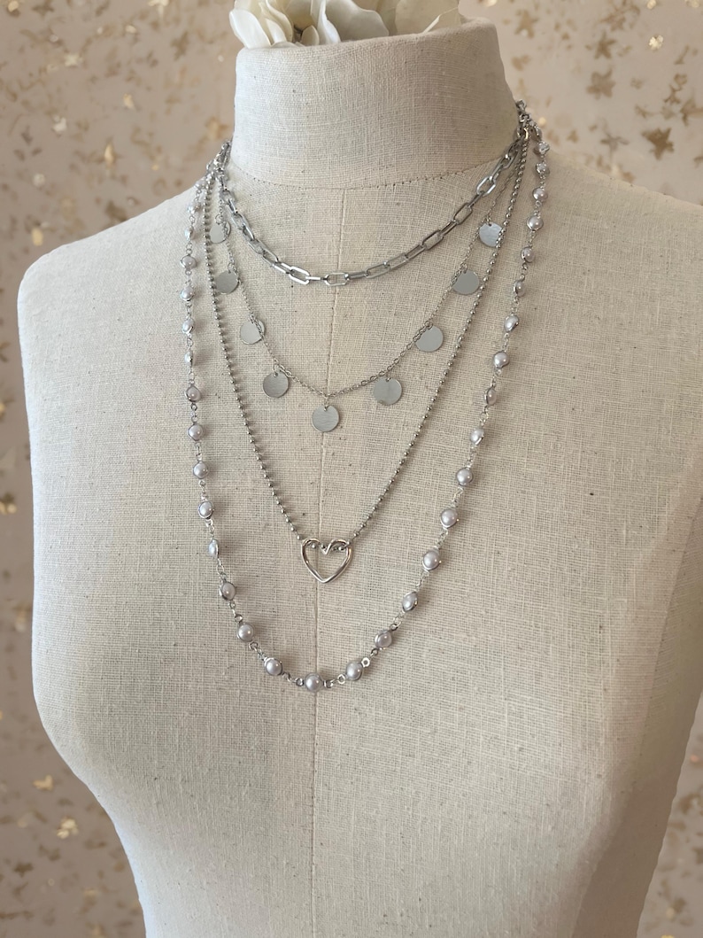 May include: A layered silver-tone necklace set featuring four distinct strands. The top layer is a thick chain, the second has small circular discs, the third features a delicate heart charm, and the longest strand is adorned with small round beads.