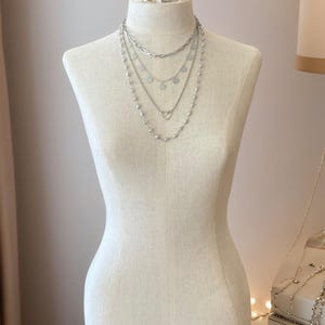 May include: A layered silver-toned necklace set featuring four distinct chains. The top chain is a thick oval link, followed by a delicate chain with small circular discs, a chain with a central heart charm, and a long chain with small beads.