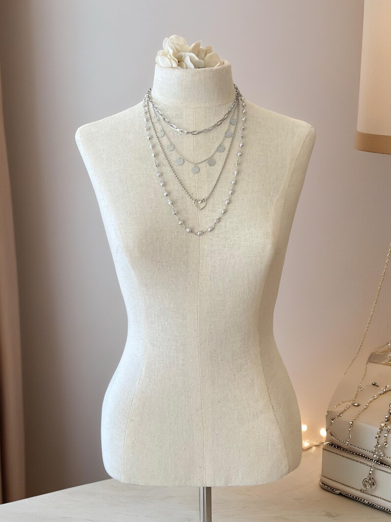 May include: A layered silver-toned necklace set featuring four distinct chains. The top chain is a thick oval link, followed by a delicate chain with small circular discs, a chain with a central heart charm, and a long chain with small beads.