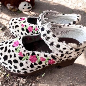 May include: A pair of children's Mary Jane shoes with a spotted pattern in black and white. The shoes feature pink floral embroidery and a brown sole. The shoes are made of leather and have a strap closure.