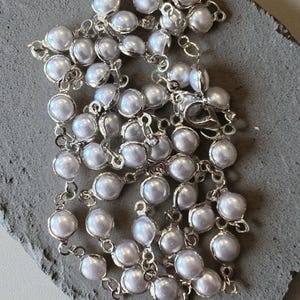 May include: A silver-toned chain necklace featuring a series of round, iridescent white faux pearls, each individually set in a metallic bezel. The necklace includes a lobster claw clasp and is arranged in a coiled, layered pattern.