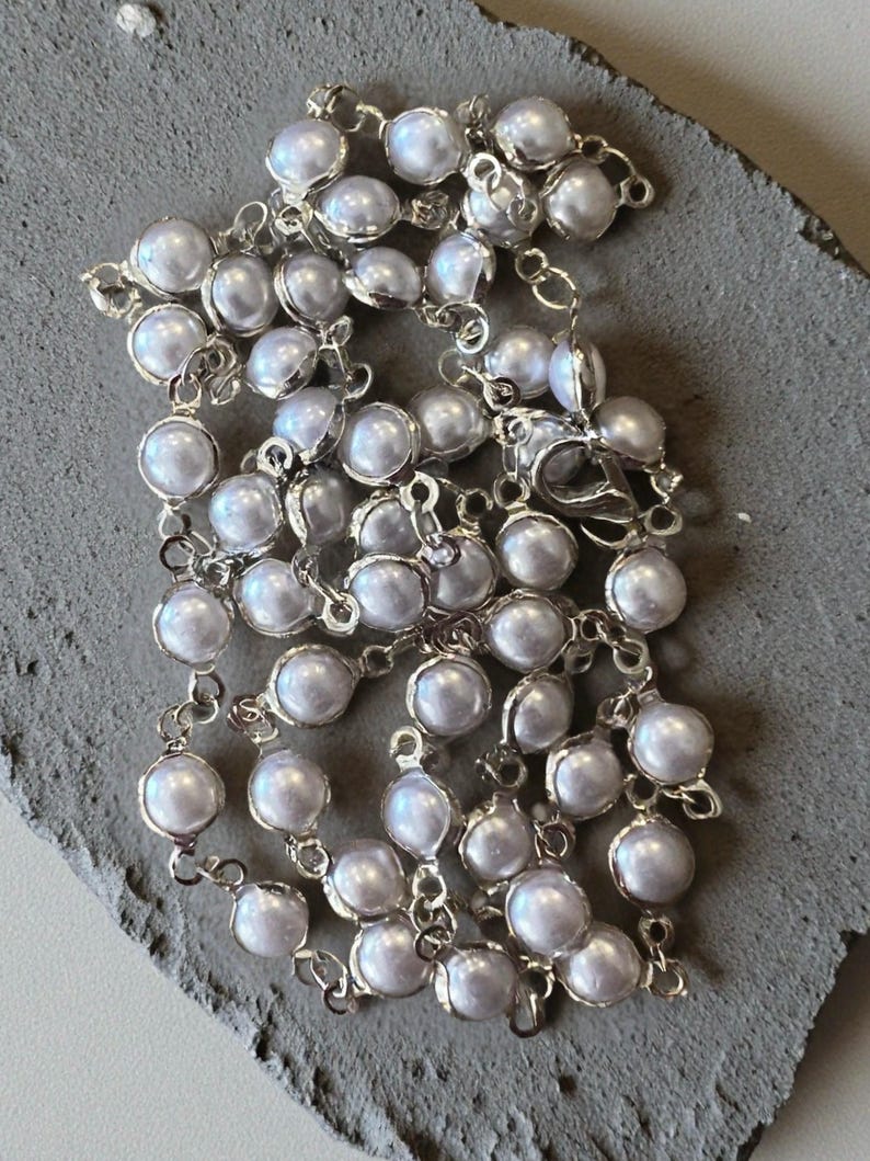 May include: A silver-toned chain necklace featuring a series of round, iridescent white faux pearls, each individually set in a metallic bezel. The necklace includes a lobster claw clasp and is arranged in a coiled, layered pattern.