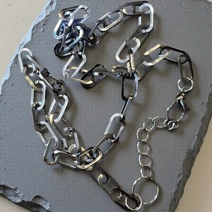 May include: A silver-toned chain necklace featuring elongated, rectangular links with a polished, reflective finish. The necklace includes a smaller circular link chain section at one end, terminating in a lobster claw clasp for secure fastening.