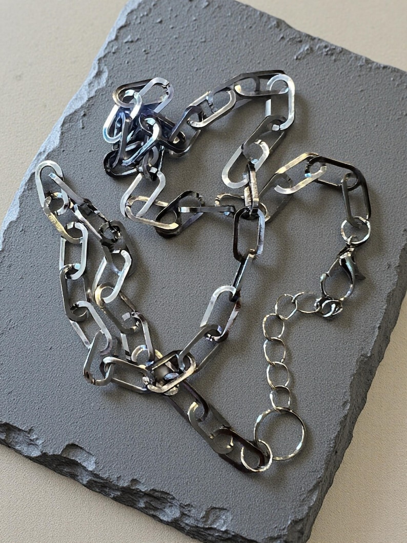 May include: A silver-toned chain necklace featuring elongated, rectangular links with a polished, reflective finish. The necklace includes a smaller circular link chain section at one end, terminating in a lobster claw clasp for secure fastening.