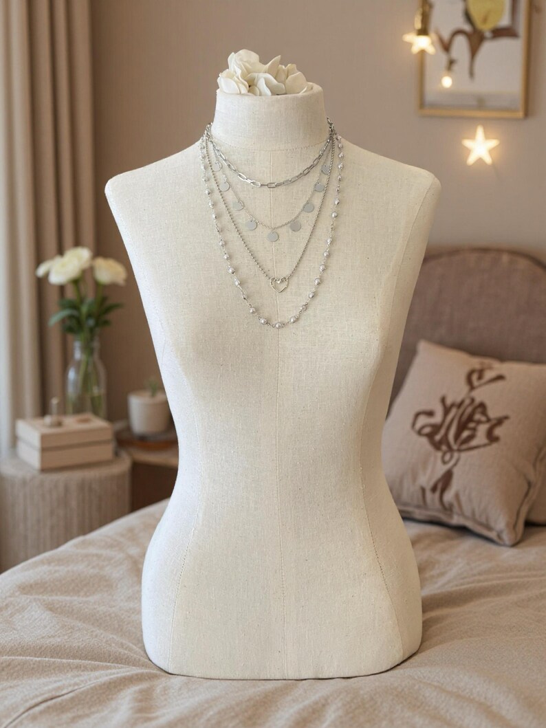 May include: A multi-layered silver-toned necklace set displayed on a beige fabric mannequin. The necklace features four distinct strands, including a thick chain, a delicate chain with small circular discs, a heart-shaped pendant, and a beaded strand.