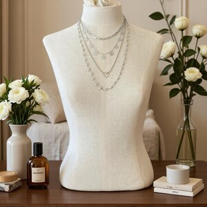 May include: A layered silver-tone necklace set displayed on a beige fabric mannequin. The set features four distinct chains: a thick oval link chain, a chain with small circular discs, a chain with a heart-shaped pendant, and a beaded chain.