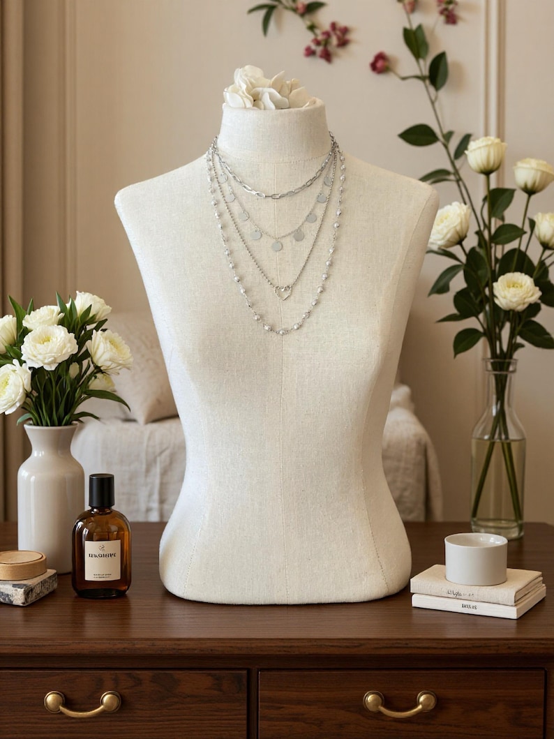 May include: A layered silver-tone necklace set displayed on a beige fabric mannequin. The set features four distinct chains: a thick oval link chain, a chain with small circular discs, a chain with a heart-shaped pendant, and a beaded chain.