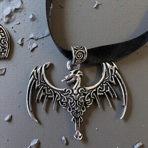 May include: A silver-toned metal dragon pendant featuring intricate knotwork detailing on its wings and body. The pendant is suspended from a smooth black ribbon necklace, which passes through a decorative silver-toned bail with a spiral pattern.
