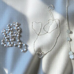 May include: A collection of three silver-toned necklaces. One necklace features a string of small white pearls, another is a delicate chain with a central open heart pendant, and the third is a thin chain accented with small, flat, circular silver discs.