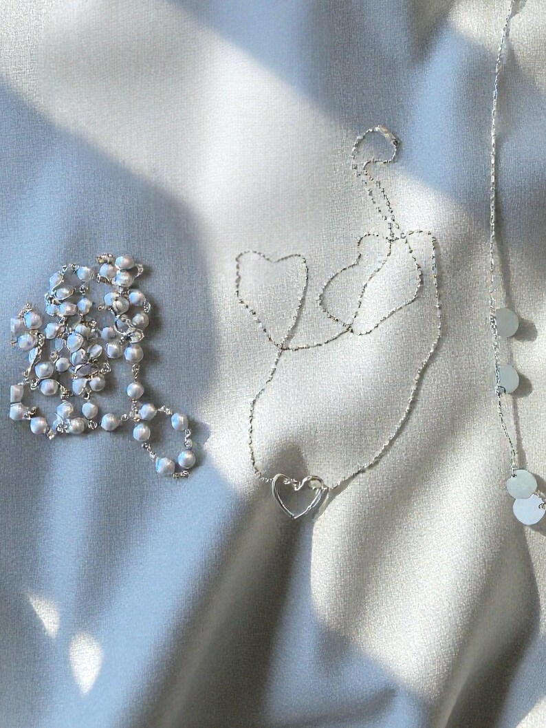 May include: A collection of three silver-toned necklaces. One necklace features a string of small white pearls, another is a delicate chain with a central open heart pendant, and the third is a thin chain accented with small, flat, circular silver discs.