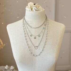 May include: A layered silver-toned necklace set featuring four distinct strands. The top layer is a thick chain, followed by a delicate chain with small circular discs, a chain with a central heart charm, and a long strand of small silver beads.