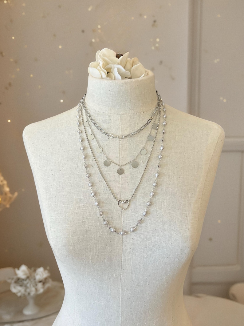May include: A layered silver-toned necklace set featuring four distinct strands. The top layer is a thick chain, followed by a delicate chain with small circular discs, a chain with a central heart charm, and a long strand of small silver beads.