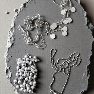 May include: Three silver-toned necklaces: one featuring a thick chain with small circular charms, one composed of small white spherical beads, and one delicate ball chain necklace with a single open heart-shaped pendant at the center.