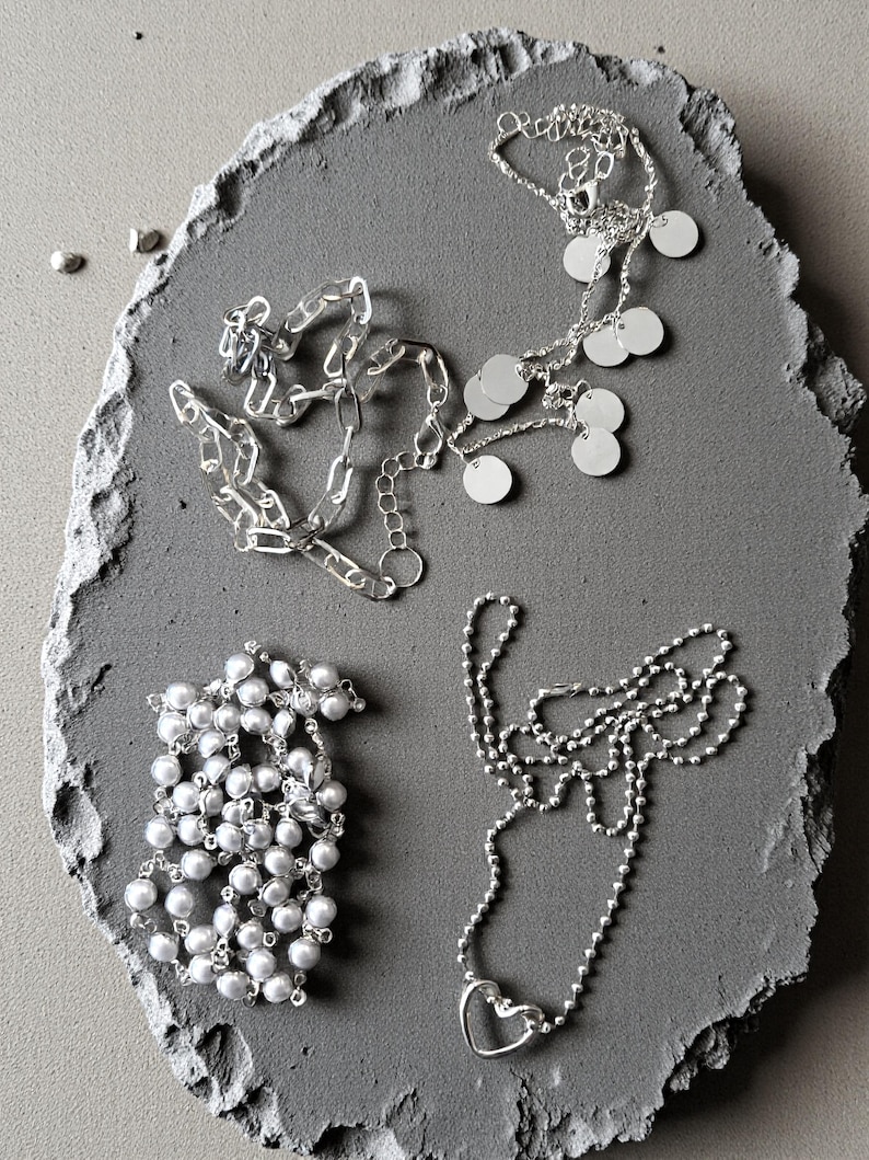 May include: Three silver-toned necklaces: one featuring a thick chain with small circular charms, one composed of small white spherical beads, and one delicate ball chain necklace with a single open heart-shaped pendant at the center.