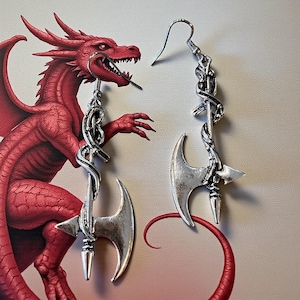 May include: Silver-toned axe earrings with a decorative chain design. The earrings feature a curved blade and a pointed tip. The background includes a red dragon illustration.