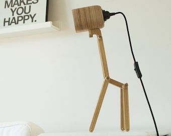 Op de afbeelding: Een houten tafellamp in de vorm van een giraf met een lichtbruine houtnerfafwerking, een cilindrische kop en scharnierende poten. De lamp is voorzien van een zwart netsnoer met een schakelaar, wat zorgt voor een functionele verlichtingsoplossing.