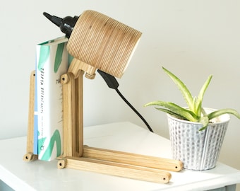 Op de afbeelding: Een lichtgekleurde houten bureaulamp met een verstelbaar scharnierend frame en een cilindrische kap. De lampvoet bevat een verticale gleuf die een boek met een witte kaft en groene abstracte markeringen vasthoudt. Een zwart netsnoer is bevestigd.
