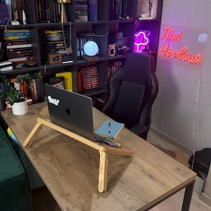 May include: A wooden laptop stand with a laptop on top, placed on a large wooden desk. A notebook and pen are next to the laptop. The desk is in front of a bookshelf filled with books and decorative items. A neon sign is visible in the background.