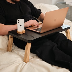 May include: A black laptop desk with wooden legs and a phone stand. A rose gold laptop and a white smartphone are on the desk. The desk is on a bed with a white comforter.