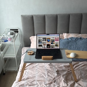 May include: A gray laptop desk with wooden legs is set up on a bed with pink and white patterned bedding. A black laptop is on the desk, along with a wooden mouse pad and a white mouse. A small wooden block and a wooden phone stand are also on the desk.