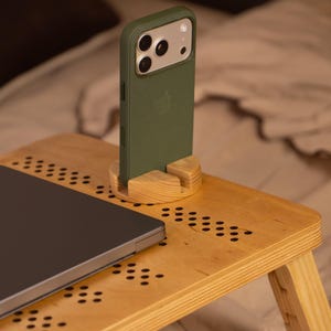 May include: A wooden laptop desk with a built-in phone stand. A green phone case is in the stand. A laptop is on the desk. The desk has a light wood finish with decorative holes. The desk is on a bed.