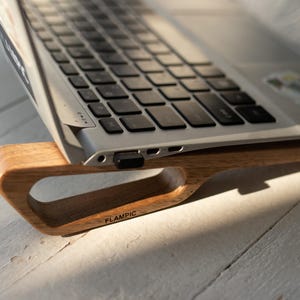Peut inclure: Un ordinateur portable argenté posé sur un support en bois brun clair avec une découpe triangulaire. Le mot FLAMPIC est imprimé en noir sur le côté du support en bois. L'ordinateur présente un clavier noir et un petit autocollant sur le repose-poignet.