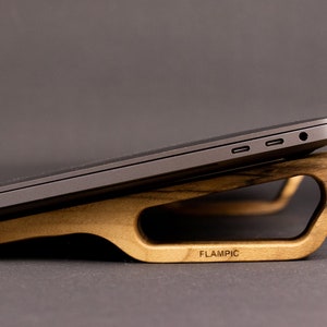 Può includere: Un portatile grigio scuro appoggiato su un supporto in legno marrone chiaro dal design liscio e curvo. Il supporto presenta un centro ritagliato e la parola FLAMPIC incisa sul lato. Il portatile è posizionato ad angolo, mostrando due porte USB-C.