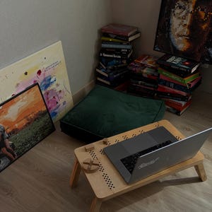 May include: A cozy workspace setup featuring a wooden laptop desk with a laptop, a green cushion, and a stack of books. Artwork and a pixelated portrait hang on the wall, creating a comfortable reading or work environment.