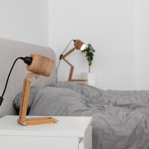 May include: A table lamp constructed from layered light-colored wood with a cylindrical head and an adjustable arm featuring metal hardware. The lamp has a black power cord and is positioned on a white surface, with a second identical lamp visible.